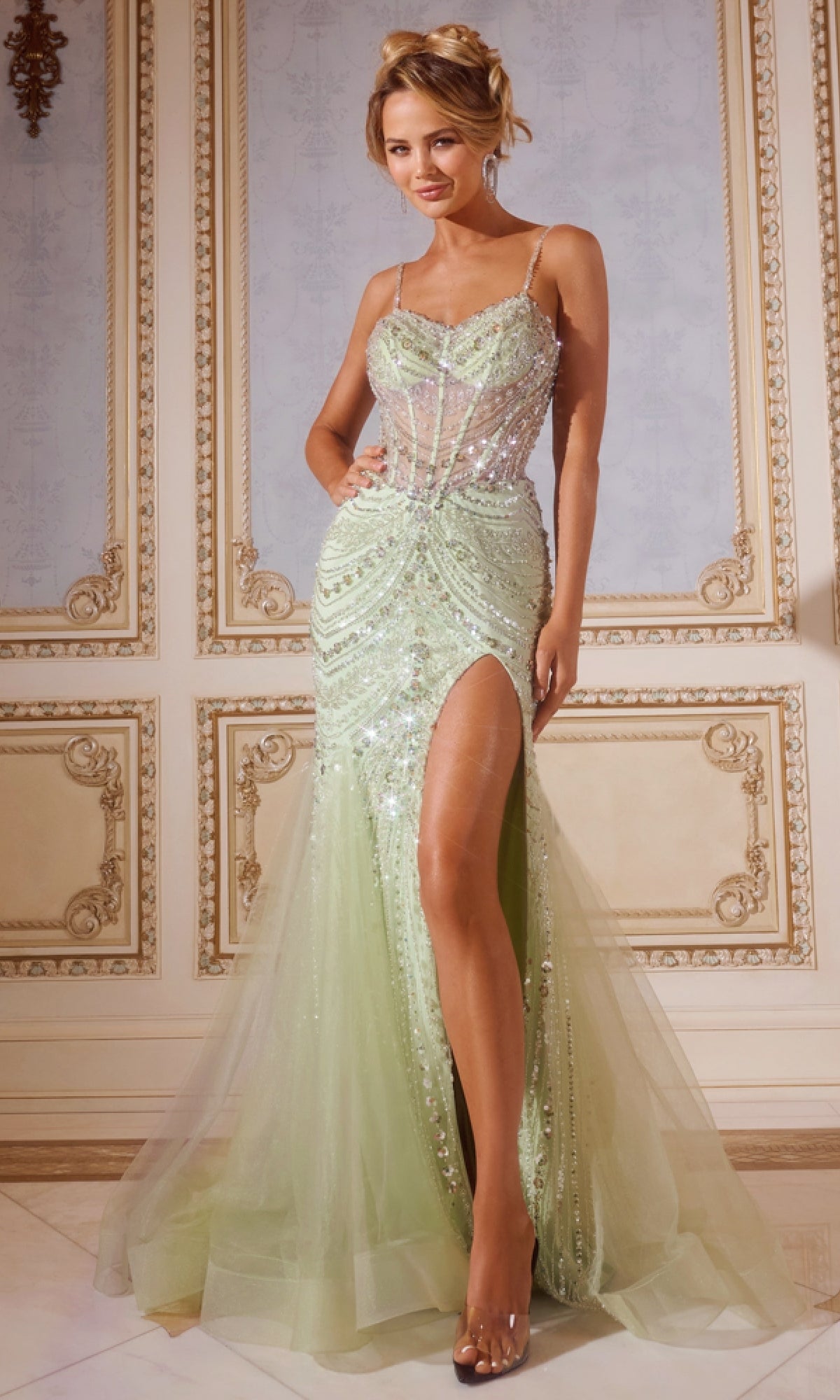 A woman wears the Jovani 43189 Long Prom Dress—a light green, sequined mermaid gown with sparkly embellishments and a thigh-high slit—while standing in an ornate room with cream-colored walls and decorative molding.